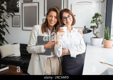 Portrait of two confident female colleagues smiling at camera holding digital tablet and cup of coffee posing in office Stock Photo