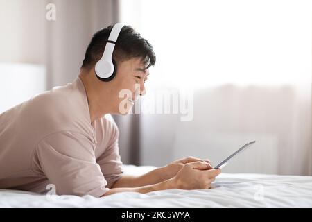 Happy asian man using digital tablet and headphones in bed Stock Photo