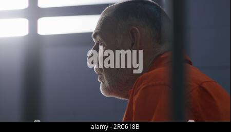 Elderly criminal in orange uniform sits on prison bed and dreams about freedom. Prisoner serves imprisonment term in jail cell. Gloomy inmate in detention center or correctional facility. Stock Photo