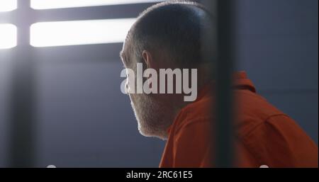 Elderly criminal in orange uniform sits on prison bed and dreams about freedom. Prisoner serves imprisonment term in jail cell. Gloomy inmate in detention center or correctional facility. Stock Photo