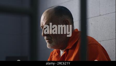 Elderly criminal in orange uniform sits on prison bed and dreams about freedom. Prisoner serves imprisonment term in jail cell. Gloomy inmate in detention center or correctional facility. Stock Photo