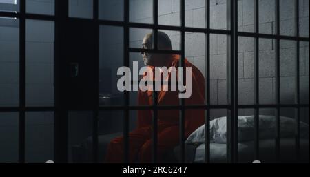 Elderly criminal in orange uniform sits on jail bed and talks with cellmate. Depressed prisoners serve imprisonment term in prison cell. Guilty inmates in detention center or correctional facility. Stock Photo