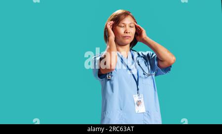 Fatigued nurse suffering from migraine pain after tiresome day at work. Hospital worker isolated over studio background affected by distressing headache during demanding job shift, close up Stock Photo