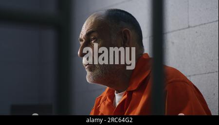 Elderly criminal in orange uniform sits on prison bed and dreams about freedom. Prisoner serves imprisonment term in jail cell. Gloomy inmate in detention center or correctional facility. Stock Photo