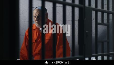 Elderly criminal in orange uniform sits on prison bed and thinks about freedom. Prisoner serves imprisonment term in jail cell. Guilty inmate in detention center or correctional facility. Stock Photo