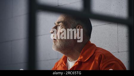 Elderly criminal in orange uniform sits on prison bed and dreams about freedom. Prisoner serves imprisonment term in jail cell. Gloomy inmate in detention center or correctional facility. Stock Photo