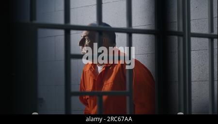 Elderly criminal in orange uniform sits on prison bed and thinks about freedom. Prisoner serves imprisonment term in jail cell. Guilty inmate in detention center or correctional facility. Stock Photo