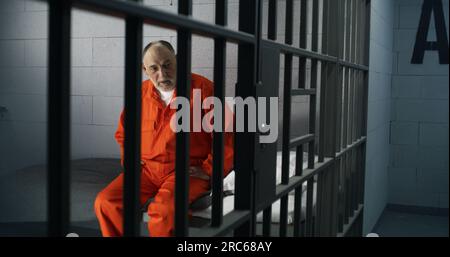 Elderly criminal in orange uniform sits on prison bed and thinks about freedom. Prisoner serves imprisonment term in jail cell. Guilty inmate in detention center or correctional facility. Stock Photo