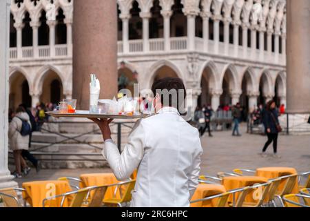 Venice, Italy - April 2, 2022: Young waiter in white suits and wearing a face mask working at a terrace cafe in San Marco Square. Stock Photo