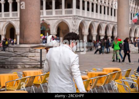 Venice, Italy - April 2, 2022: Young waiter in white suits and wearing a face mask working at a terrace cafe in San Marco Square. Stock Photo