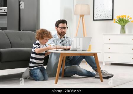 Man working remotely at home. Father using laptop while his son drawing at desk Stock Photo