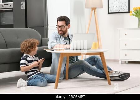 Man working remotely at home. Father using laptop at desk while his son playing with smartphone in living room Stock Photo