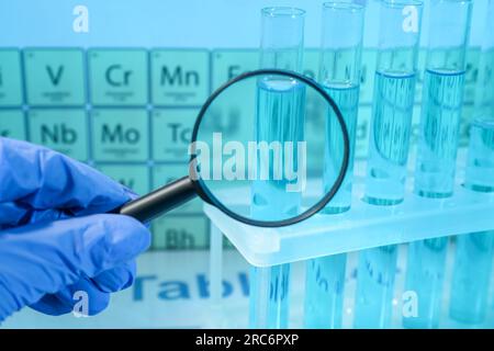 Scientist with magnifying glass examining test tube against periodic table of elements in laboratory, closeup. Color tone effect Stock Photo