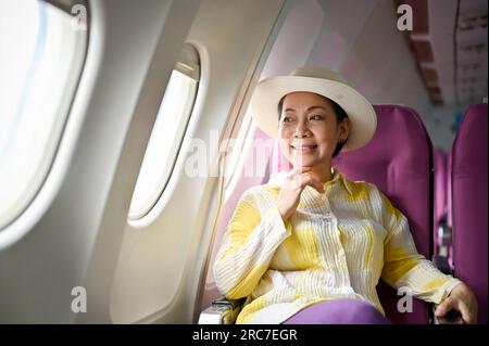 A portrait of an attractive and happy senior Asian female passenger in a summer outfit sits at her window seat on a plane going for her summer vacatio Stock Photo