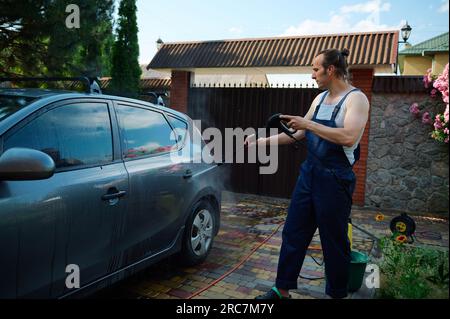 Handsome muscular attractive young adult man in blue work overalls, directing a high pressure water jet on his car while cleaning it outdoors, using a Stock Photo