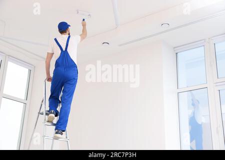 Worker in uniform painting ceiling with roller on stepladder indoors, back view. Space for text Stock Photo