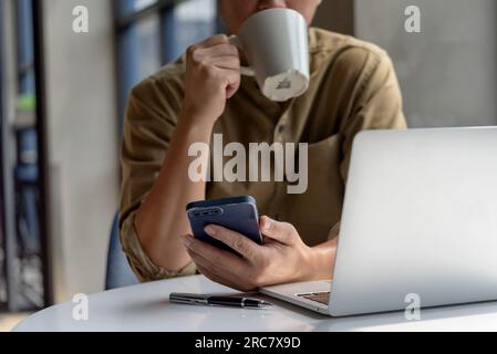 man asian drinking coffee and looking mobile phone computer laptop and at desk. Business technology digital checking emails or browsing social media w Stock Photo
