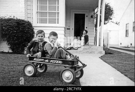 United States:  c. 1951. Two brothers sitting in their new Radio Flyer wagon while their mother looks on from the front steps. Stock Photo