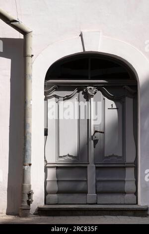 Old gray weathered decorated semi-circular door in a plastered white wall with a drainpipe next to it in Riga, Latvia Stock Photo