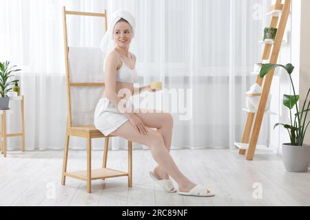 Beautiful young woman applying body cream onto leg indoors Stock Photo