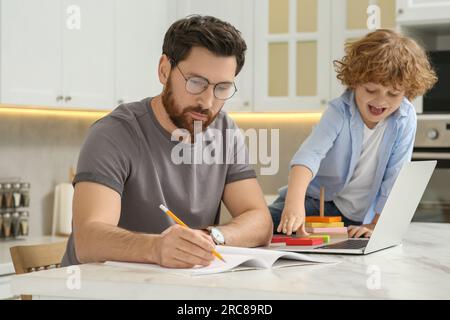 Father working remotely while his son playing with toys at home. Man writing in notebook at desk Stock Photo