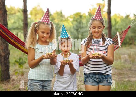 Children holding birthday cakes with burning candles. Kids party decoration and food. Boy and girls celebrating birthday in the garden with hammock. K Stock Photo