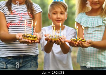 Children holding birthday cakes with burning candles. Kids party decoration and food. Boy and girls celebrating birthday in the garden with hammock. K Stock Photo