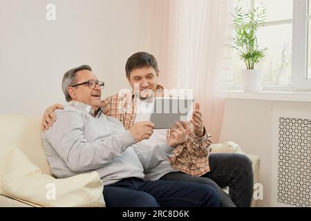teacher son. Smiling young male glad to explain work online in app on laptop to beloved mature dad. Elderly father and adult kid use tablet for web sh Stock Photo