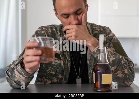 Unhappy man in camouflage uniform sitting at arm chair over white studio background, touching his head, soldier suffering from posttraumatic stress Stock Photo