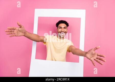 Photo of funny cheerful candid guy wear stylish yellow t-shirt look from frame stretching hands to you isolated on pink color background Stock Photo