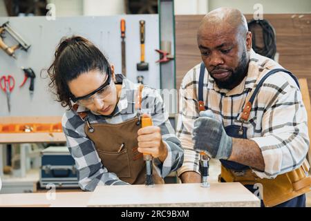strong woman worker. builder team work. joiner in wood workshop. lady carpenter staff helping man working making furniture. hard work fix wooden board Stock Photo