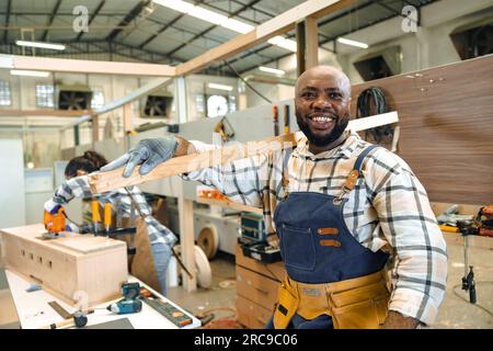 happy carpenter wood enjoy working in large wood workshop. smiling black joiner carrying pile wood making furniture Stock Photo