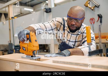 black african male carpenter joiner using Jig Saw wood panel cutter to cut making furniture panel in workshop Stock Photo