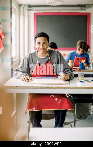 Portrait of smiling male teenage student sitting at desk during art class in high school Stock Photo