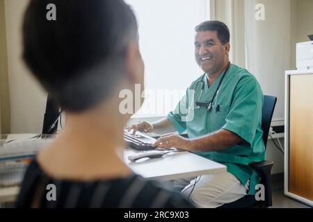 Happy male doctor looking at patient while using computer in clinic Stock Photo