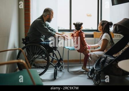 Mother and daughter looking through window by man in wheelchair at hospital Stock Photo