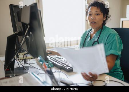 Female doctor examining medical record while using computer at desk in office Stock Photo
