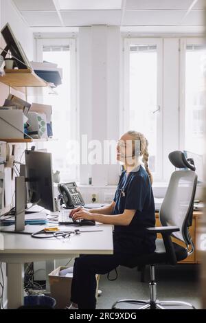 Side view of happy female healthcare worker sitting and using computer at desk in clinic Stock Photo