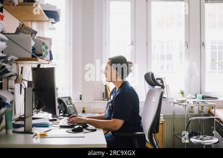 Side view of focused female healthcare worker using computer while sitting on desk in clinic Stock Photo