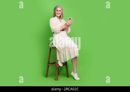 Full size photo of attractive old woman sit wooden chair comment influencer wear trendy white clothes isolated on green color background Stock Photo