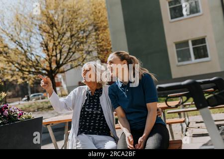 Senior woman gesturing while sitting with female caregiver on bench Stock Photo