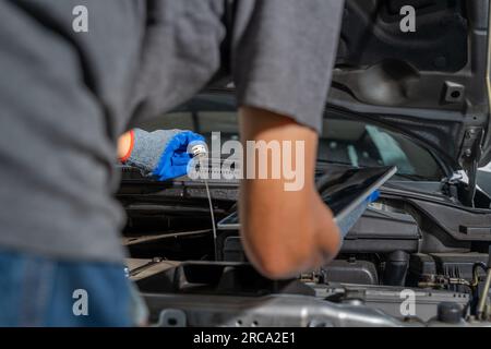 A male car mechanic checking car motor oil and holding a computer tablet while working on repairing the car engine at his car repair garage Stock Photo