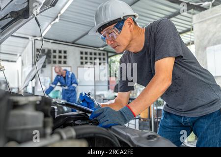 An Asian male car mechanic writing down notes on his notepad while working on repairing a car at his car repair garage, another male mechanic working Stock Photo