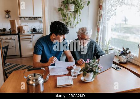 Male caregiver discussing medical reports with senior man using laptop at home Stock Photo