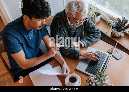 Senior man using laptop while sitting next to male caregiver explaining medical reports at home Stock Photo