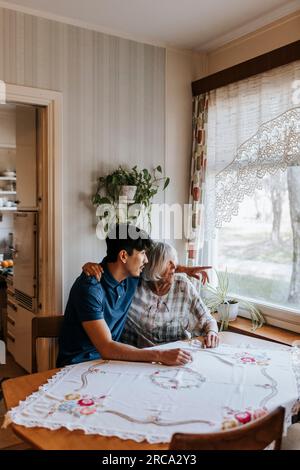 Male healthcare worker gesturing while sitting with senior woman near window at home Stock Photo