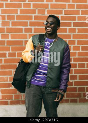 Happy young man with backpack while standing in front of brick wall Stock Photo