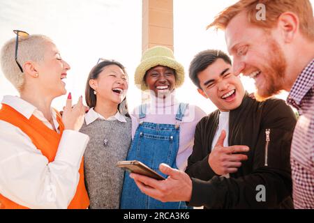 Multiracial group of young people enjoying and having fun using a cellphone and watching funny messages outside. Joyful man showing the social media to his friends on smart phone and laughing together. High quality photo Stock Photo