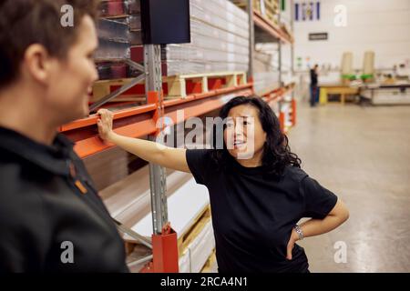 Mature female blue-collar worker discussing with colleague while standing by rack in distribution warehouse Stock Photo