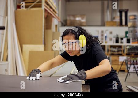 Serious female carpenter wearing protective eyewear and ear protectors while working in industry Stock Photo